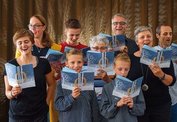 Neun Leute stehen zusammen und singen, in den Händen halten sie das Weihnachtsliederheft