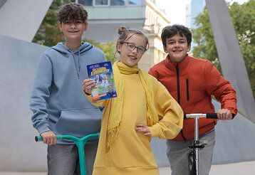 zwei Jungen mit Scootern und ein Mädchen stehen vor einem Gebäude, das Mädchen hält ein Buch in die Kamera