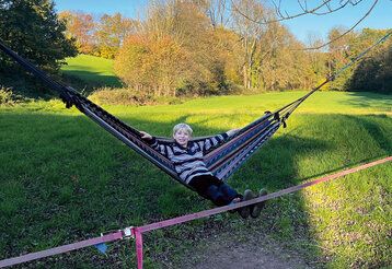 Junge auf Hängematte, im Hintergrund eine große Wiese mit Bäumen, herbstlich