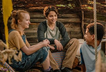 ein Mädchen und zwei Jungen sitzen in einer Holzhütte und unterhalten sich