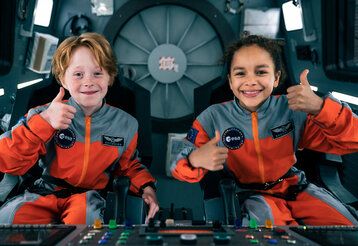 Junge und Mädchen sitzen mit Astronautenanzügen im Cockpit eines Raumschiffe und halten die Daumen hoch