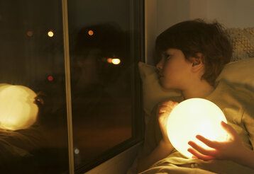 Junge liegt in dunklem Raum mit runder Leuchte im Bett und schaut aus dem Fenster