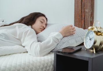 Frau im weissen Hemd liegt auf weissem Bett mit einem goldenen Wecker an der Seite