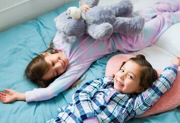 Zwei Mädchen lieben auf einem Bett, in gegengesetzten Richtungen, eine hat ein Plüschtier im Arm