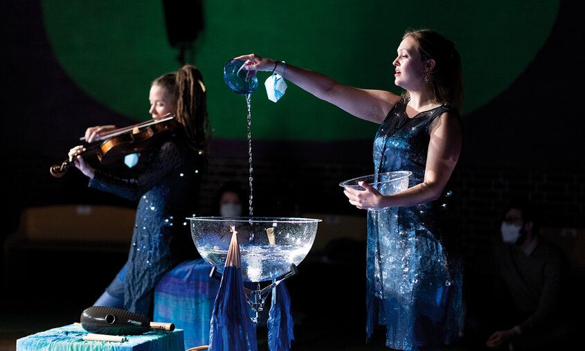 Eine Frau schüttet Wasser in eine Schale, im Hintergrund spielt eine Frau Geige