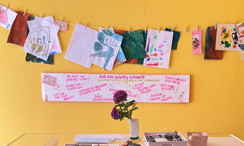 Art Bakery, Tisch mit Malutensilien und Hortensie vor gelber Wand, gespannte Leine mit Kinderbildern