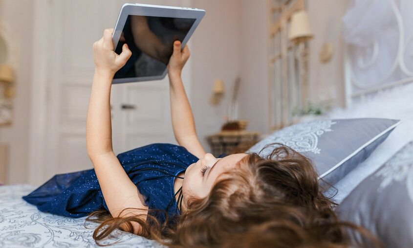 ein kleines Mädchen liegt auf dem Bett und hält ein Tablet in den Händen